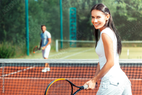 Dark-eyed wife smiling while playing tennis with husband