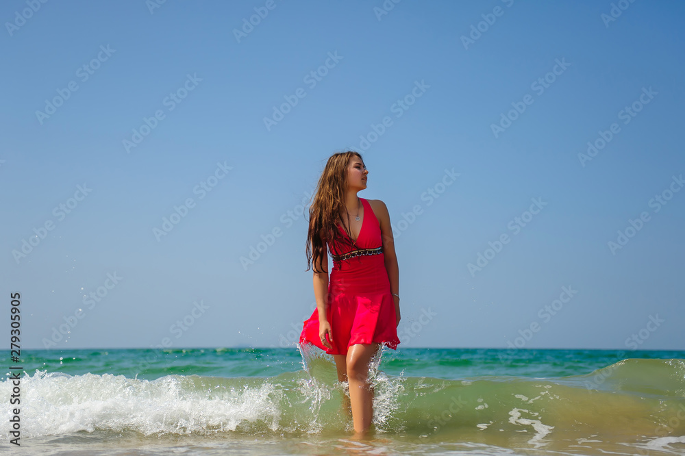 Young sexy long haired brunette in red dress standing in waves with white foam on sea background and blue sky on hot day. Beautiful woman posing in turquoise ocean water during vacation. Young sexy long haired brunette in red dress standing in waves with white foam on sea background and blue sky on hot day. Beautiful woman posing in turquoise ocean water during vacation.