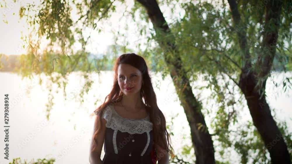 Close-up, slow motion, portrait of an attractive European girl in a dress with long brown hair by the river, lake in summer at sunset Close-up, slow motion, portrait of an attractive European girl in a dress with long brown hair by the river, lake in summer at sunset