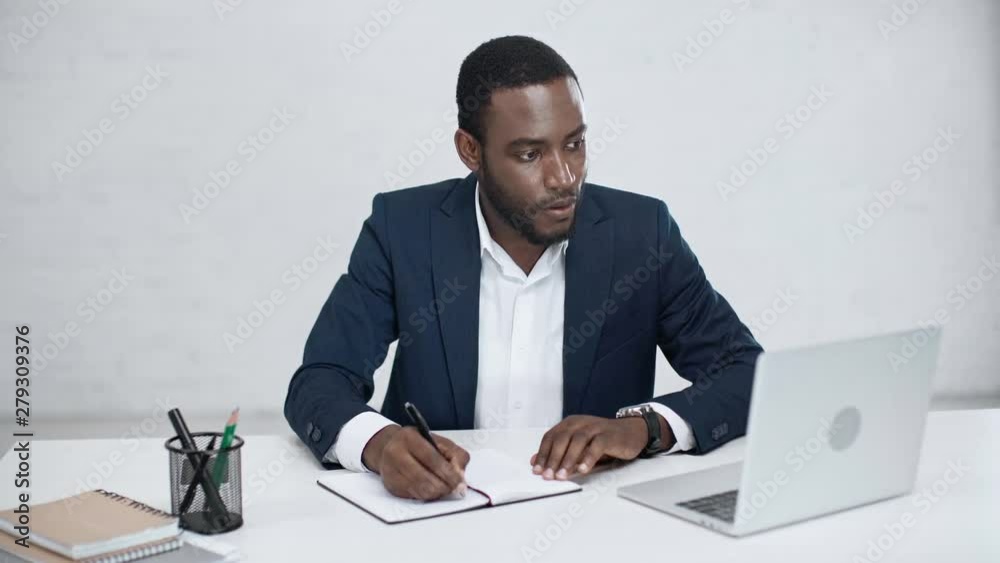 attentive african american businessman writing in notebook and using laptop while sitting at desk in office