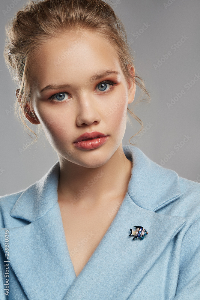 Portrait of girl with tied back fair hair, wearing sky blue coat with bright brooch in view of stripy fish on the lapel. The woman is tilting head and looking at camera, posing on gray background. Portrait of girl with tied back fair hair, wearing sky blue coat with bright brooch in view of stripy fish on the lapel. The woman is tilting head and looking at camera, posing on gray background.