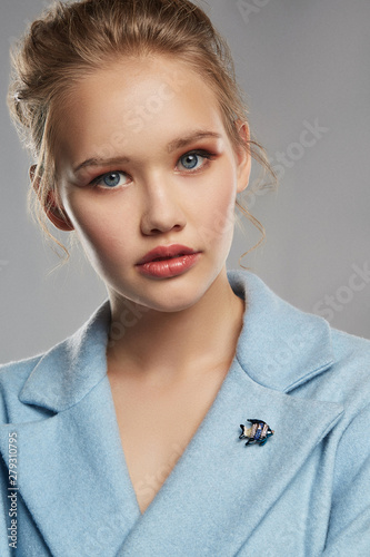 Fototapet Portrait of girl with tied back fair hair, wearing sky blue coat with bright brooch in view of stripy fish on the lapel