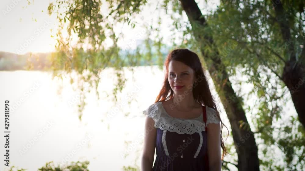 Close-up, slow motion, portrait of an attractive European girl in a dress correcting long brown hair by the river, lake in summer at sunset Close-up, slow motion, portrait of an attractive European girl in a dress correcting long brown hair by the river, lake in summer at sunset