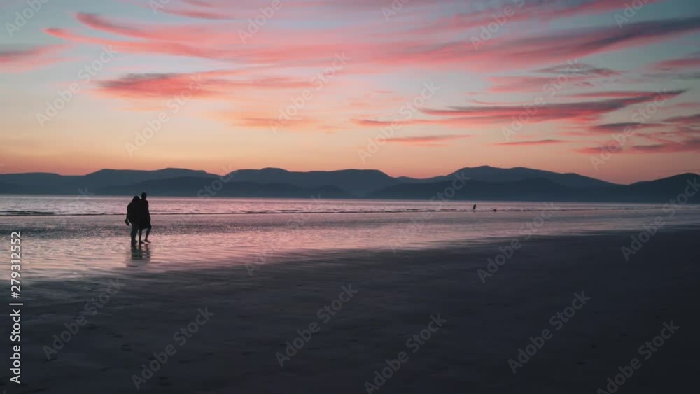 silhouettes of man and woman walking along the Beach Rossbeigh during the majestic sunset. Purple sky and mountains in the background silhouettes of man and woman walking along the Beach Rossbeigh during the majestic sunset. Purple sky and mountains in the background