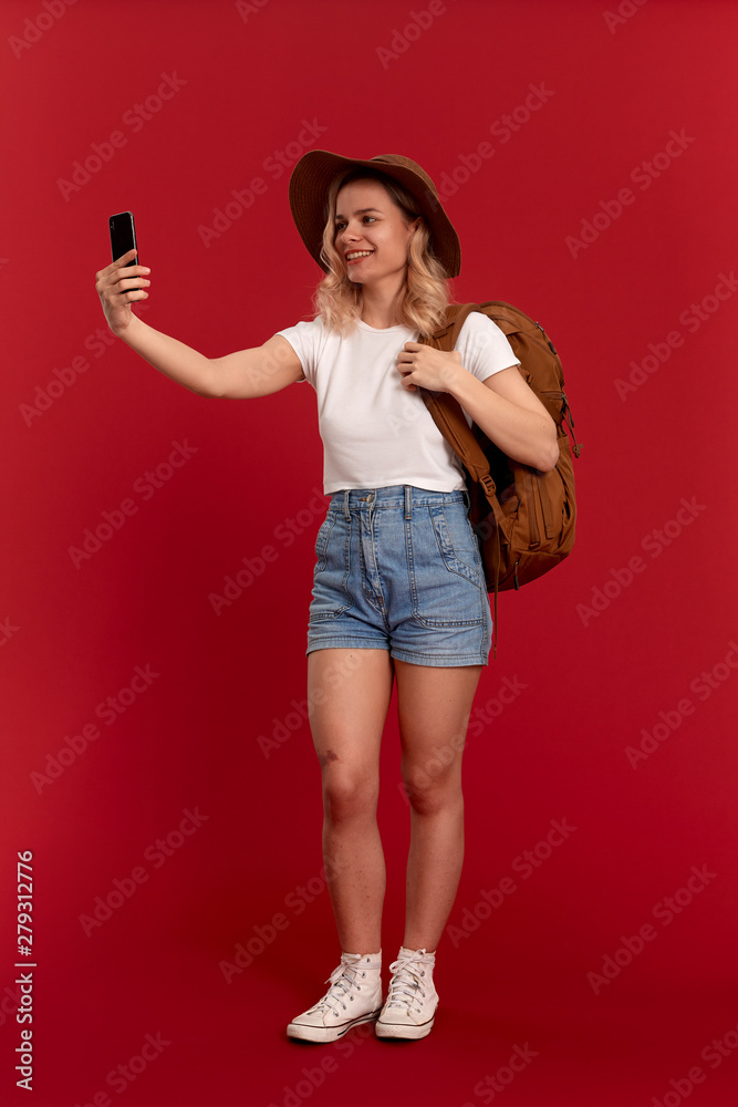 Naklejka premium Portrait of a girl with curly blond hair dressed in a sundwon hat and white t-shirt standing on a red background making selfie with mobile phone. Model travels with orange backpack.
