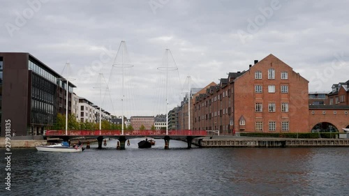 View of landmark red buildings and metal bridge over the Christianshavn Canal in Copenhagen on an overcast day with boats passing underneath it 
