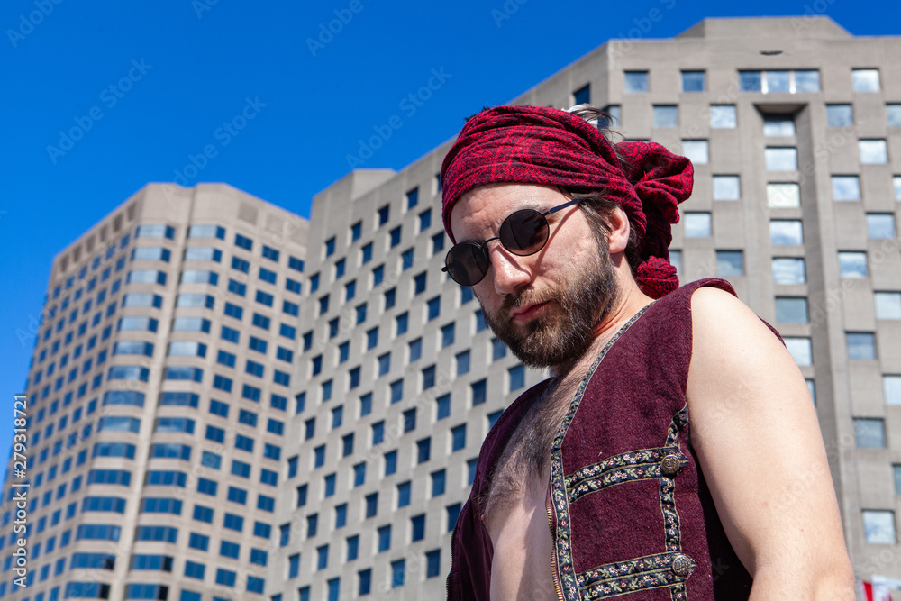 Confident man wears red bandanna in city. A low angle view of a bare chested Caucasian male wearing red headscarf and sunglasses by a commercial building. Artistic person seeks inspiration in city.