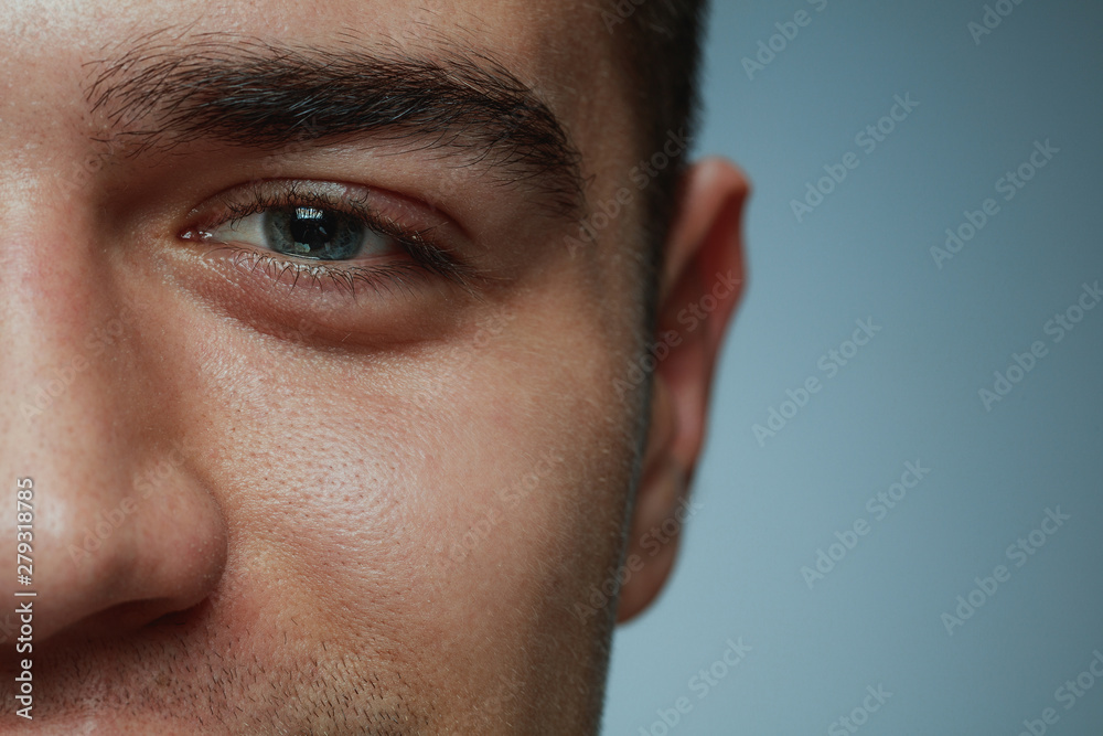 Close-up portrait of young man isolated on grey studio background. Caucasian male model's face and blue eye. Concept of men's health and beauty, self-care, body and skin care, medicine or phycology.