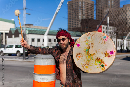 Spiritual man holds native drum downtown. A peaceful shaman in his thirties is seen shirtless, holding a sacred drum and stick on a busy city street, blurred commercial buildings in background.
