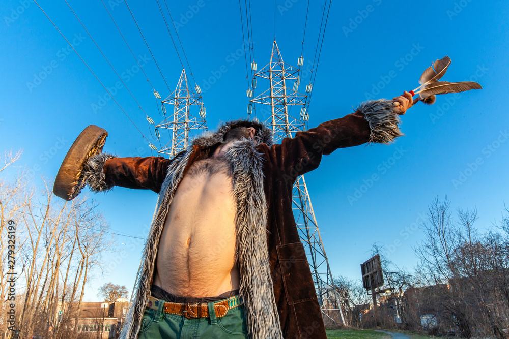 Naklejka premium Sacred man beneath electricity towers. A front view of a spiritual man as he meditates, standing beneath electric pylons in a city. Low angled shot of bare chested Caucasian bohemian.