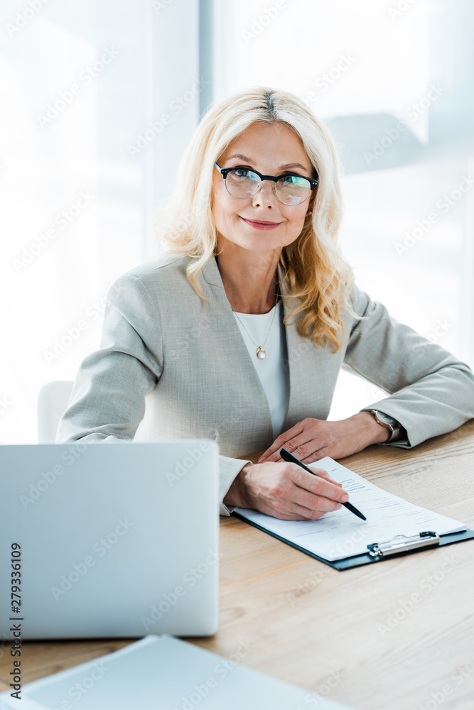 happy blonde woman in glasses looking at camera while holding pen near clipboard and laptop happy blonde woman in glasses looking at camera while holding pen near clipboard and laptop