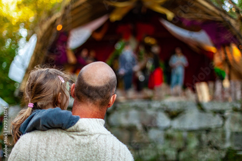 Fusion of cultural & modern music event. A bald headed father is viewed from behind, holding his his little girl as they watch a live band perform at a family music festival, with copy space.