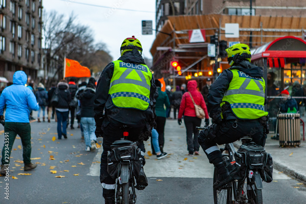 Obraz premium Environmental activists march in city. Two policemen are seen from the rear, riding bicycles during a demonstration by environmentalists, protestors are seen in the background on an urban street