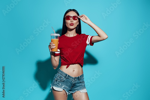 Juice cocktail. Attractive young Asian woman puckering and adjusting eyewear while standing against blue background