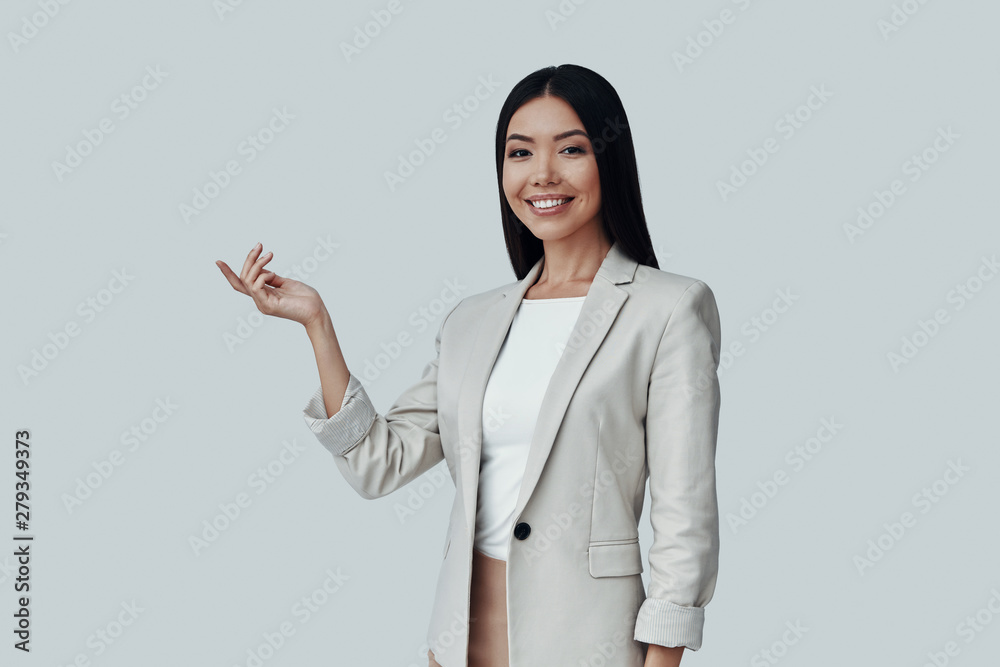 Take a look! Attractive young Asian woman looking at camera and pointing copy space while standing against grey background Take a look! Attractive young Asian woman looking at camera and pointing copy space while standing against grey background