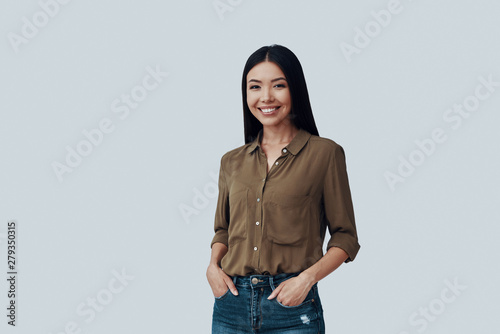Young and beautiful. Attractive young Asian woman looking at camera and smiling while standing against grey background