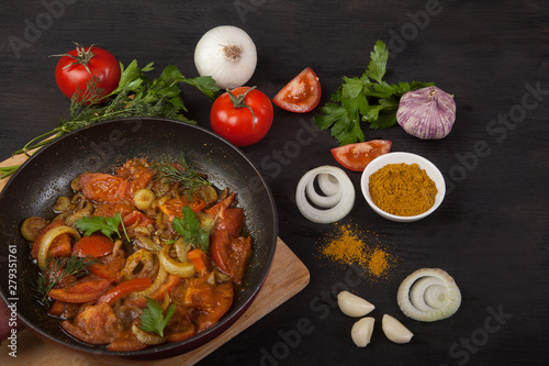 Roasted tomatoes with onions in a pan and fresh vegetables on a black background. Top view