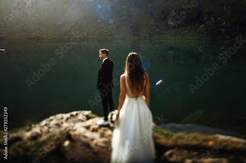 Amazing caucasian groom wainting his bride while looking at the epic view of mountains against sunrise near a lake. photo