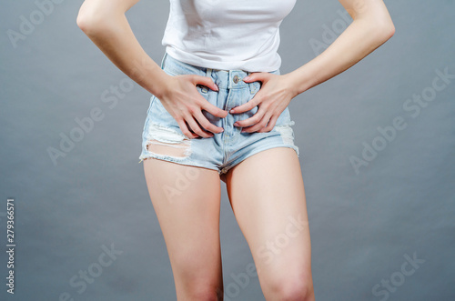 Woman with slender legs dressed in short denim shorts presses her hands to the lower abdomen, experiencing pain in the crotch.