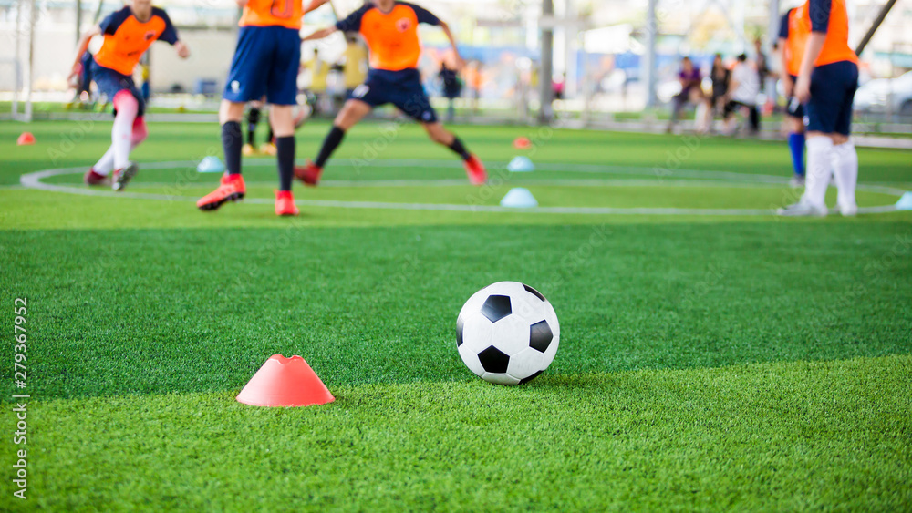 football on green artificial turf with blurry soccer team training, blurry kid soccer player jogging between marker cones and control ball with soccer equipment in soccer academy.