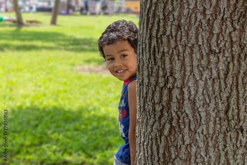 The small boy pears from behind a large tree with a smile looking at the camera. Beautiful summer day at the park for all to enjoy.