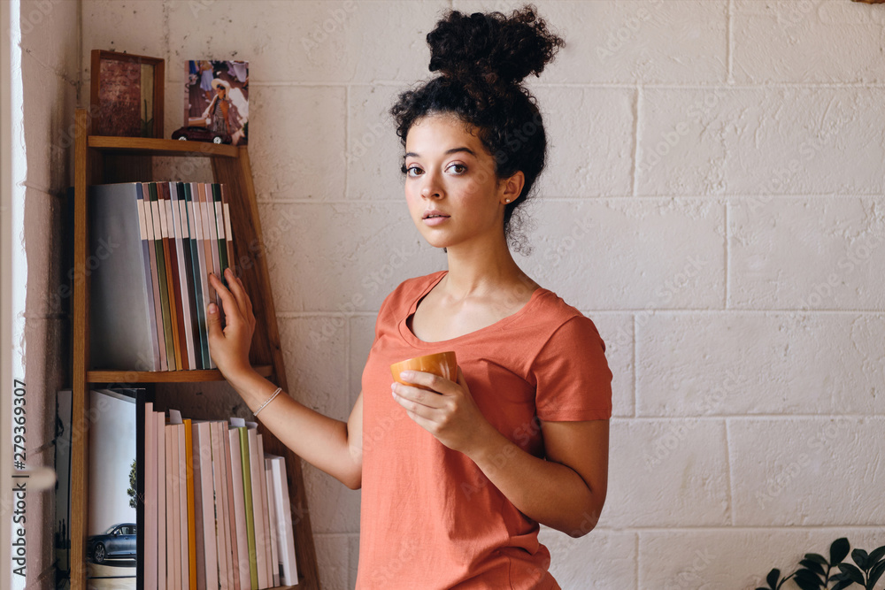 Young attractive woman with dark curly hair in T-shirt holding cup of coffee in hand and thoughtfully looking in camera standing near bookshelf at cozy home Young attractive woman with dark curly hair in T-shirt holding cup of coffee in hand and thoughtfully looking in camera standing near bookshelf at cozy home