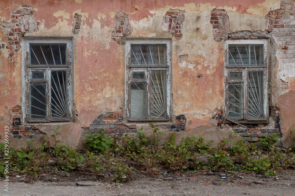Close-up view of the facade of an old abandoned house. Three windows with lattice, shabby plastered brick walls. Urban background