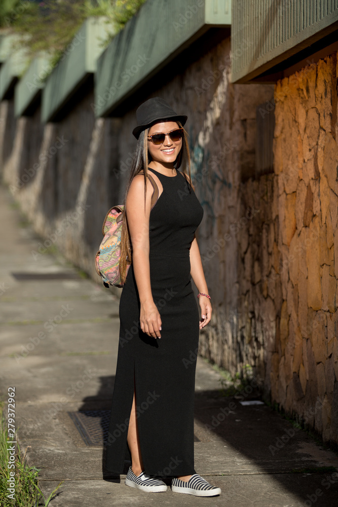 Young beautiful lady model poses in the city streets. She is wearing a black dress, sunglasses and a hat and is carrying a purse as backpack.