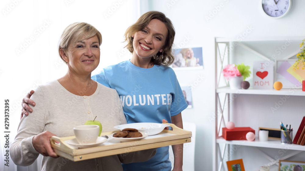 Volunteer hugging aged lady, holding tray with food, looking at camera, charity Volunteer hugging aged lady, holding tray with food, looking at camera, charity
