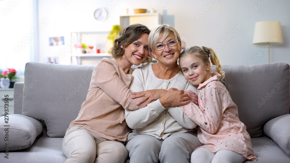 Multi-generation females hugging on home sofa smiling camera, family closeness Multi-generation females hugging on home sofa smiling camera, family closeness