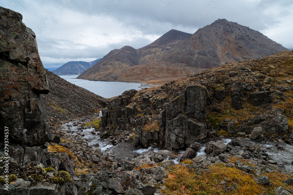 Severe northern landscape with a mountain stream in a gorge among the rocks. In the distance, a sea bay and mountains. Autumn in the Arctic. Providence Bay, Chukotka, Siberia, Far East of Russia. Severe northern landscape with a mountain stream in a gorge among the rocks. In the distance, a sea bay and mountains. Autumn in the Arctic. Providence Bay, Chukotka, Siberia, Far East of Russia.