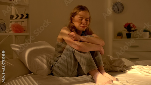 Crying young woman sitting alone in bedroom, regretting about abortion, sorrow
