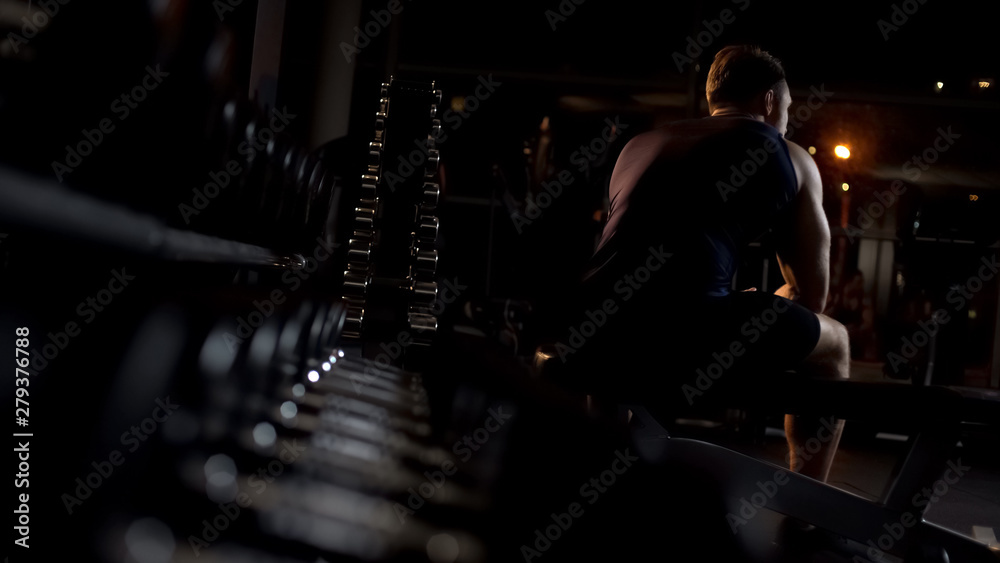 Fototapeta Male athlete tired after workout sits on bench, hunches up alone in evening gym