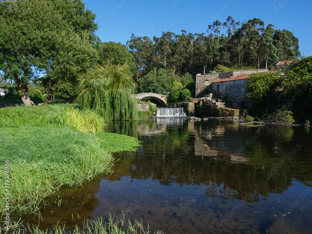 Fototapeta premium Beautiful scenic view over river Este in Portugal, on a sunny summer day with blue sky. Old stone bridge, ruin of stone house, and overhanging willow tree on bank.
