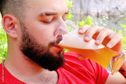 Close-up. Brutal guy with a beard in a red T-shirt drinks beer. Caucasian traveler on vacation in the woods uses alcohol. Portrait of a handsome young man.