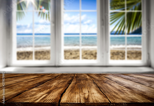 Table background with wooden top board and white window. Beautiful blurred ocean and beach view outside the window.