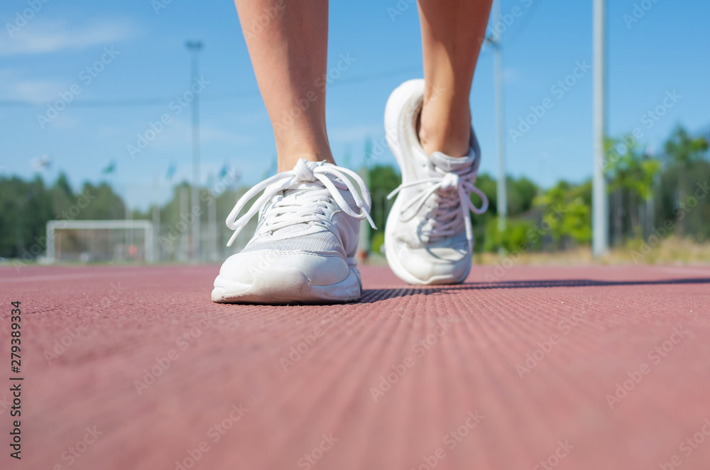 Women's legs in white sneakers are walking around the stadium. The concept of amateur sports, athletics, outdoor activities. Place for text.