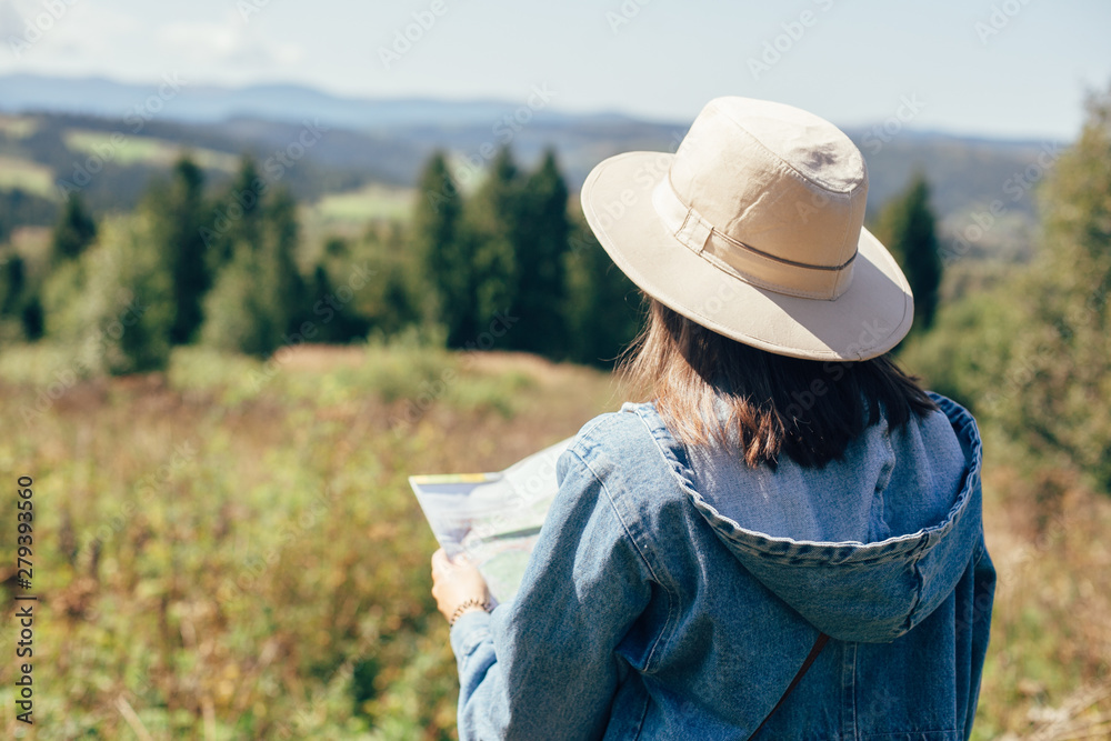 Stylish hipster girl holding map and traveling in sunny mountains. Woman in hat exploring map and hiking on top of mountain on summer vacation. Wanderlust and travel concept