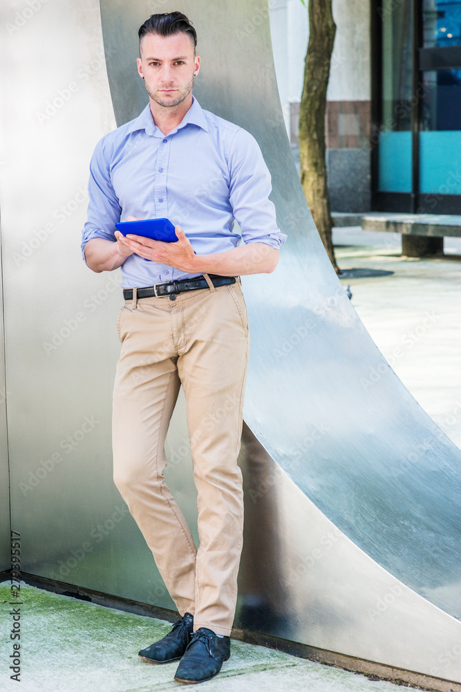 Young man reading outside in New York City, wearing light purple, long  sleeve shirt, beige pants, black shoes, standing by silver metal wall on  street, hands holding blue tablet computer, thinking.. Photos