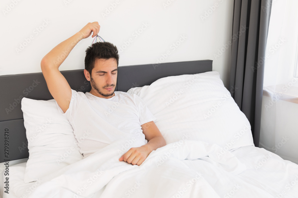 Young handsome man with a beard sits in his bed. He makes himself a head massage with a special hand massager. Young handsome man with a beard sits in his bed. He makes himself a head massage with a special hand massager.
