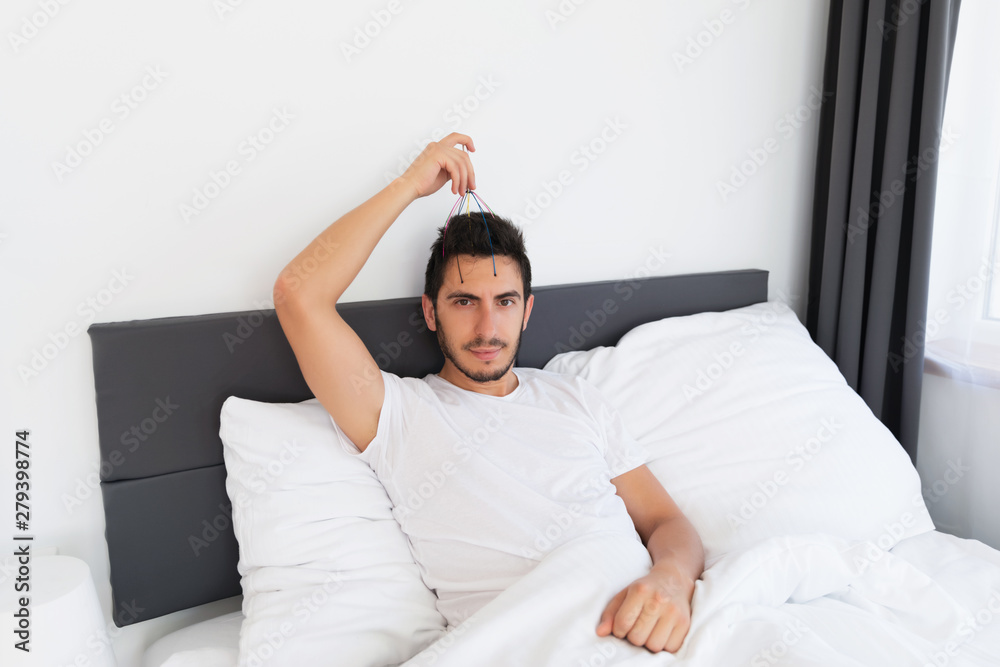 Young handsome man with a beard sits in his bed. He makes himself a head massage with a special hand massager. Young handsome man with a beard sits in his bed. He makes himself a head massage with a special hand massager.
