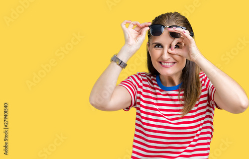 Middle age mature woman wearing sunglasses over isolated background doing ok gesture with hand smiling, eye looking through fingers with happy face.
