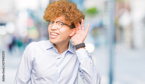 Young handsome business man with afro wearing glasses smiling with hand over ear listening an hearing to rumor or gossip. Deafness concept. © Krakenimages.com