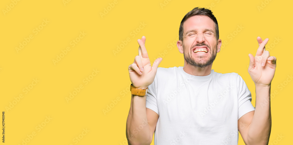 Handsome man wearing casual white t-shirt smiling crossing fingers with hope and eyes closed. Luck and superstitious concept. Handsome man wearing casual white t-shirt smiling crossing fingers with hope and eyes closed. Luck and superstitious concept.