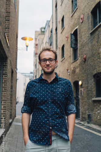 Young man wearing glasses is standing in the narrow street in the city.