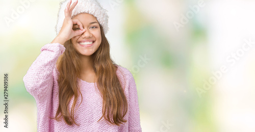 Young beautiful brunette woman wearing sweater and winter hat over isolated background doing ok gesture with hand smiling, eye looking through fingers with happy face.