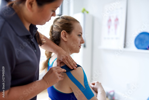 Chinese woman massage therapist applying kinesio tape to the shoulders and neck of an attractive blond client in a bright medical office