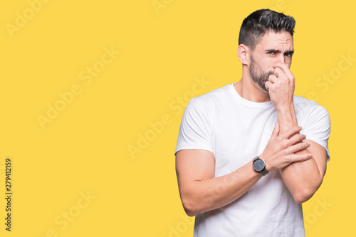 Young man wearing casual white t-shirt over isolated background smelling something stinky and disgusting, intolerable smell, holding breath with fingers on nose. Bad smells concept.