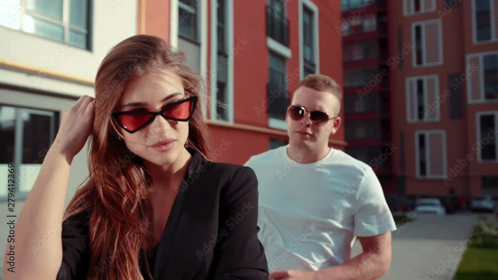Good looking young woman in trendy sunglasses stands outside, handsome strong man comes and hugs her from back, kissing in neck. Romantic mood, enjoying each other Good looking young woman in trendy sunglasses stands outside, handsome strong man comes and hugs her from back, kissing in neck. Romantic mood, enjoying each other