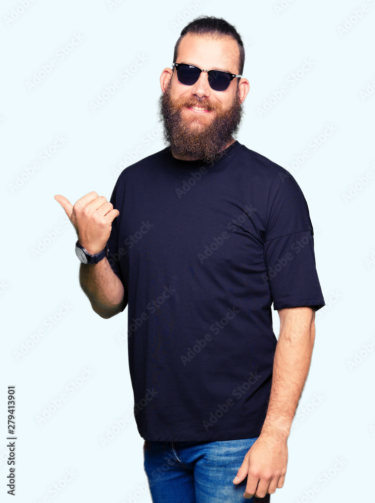 Young hipster man wearing sunglasses smiling with happy face looking and pointing to the side with thumb up. Young hipster man wearing sunglasses smiling with happy face looking and pointing to the side with thumb up.